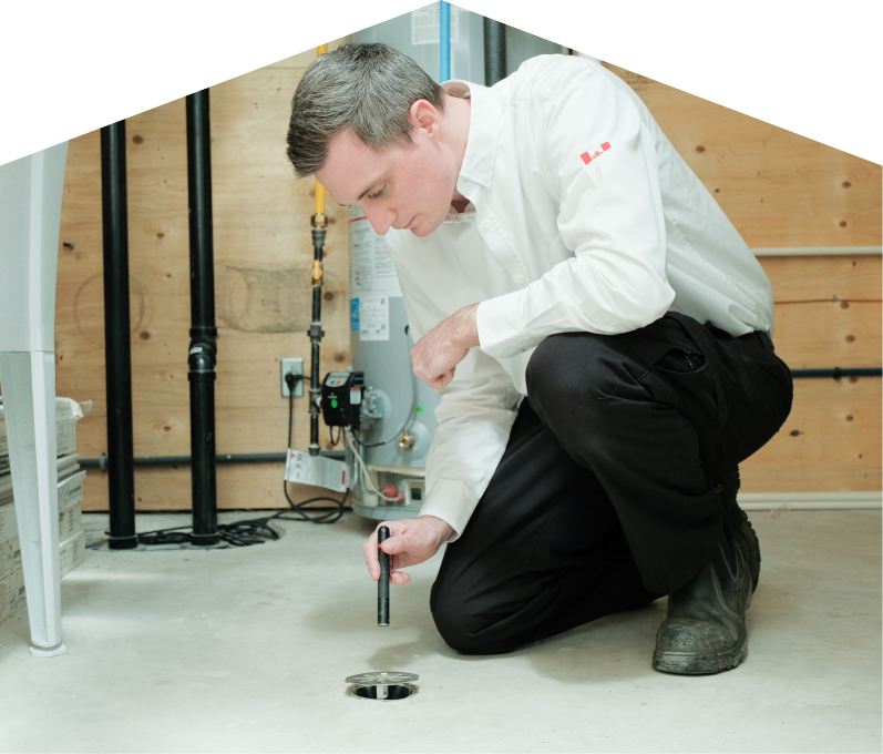 Mersey plumbing specialist inspecting a basement floor drain with a flashlight during a drain cleaning service, in a utility room with visible pipes, water heater, and wood panel walls. Mersey plumbing specialist inspecting a basement floor drain with a flashlight during a drain cleaning service, in a utility room with visible pipes, water heater, and wood panel walls.