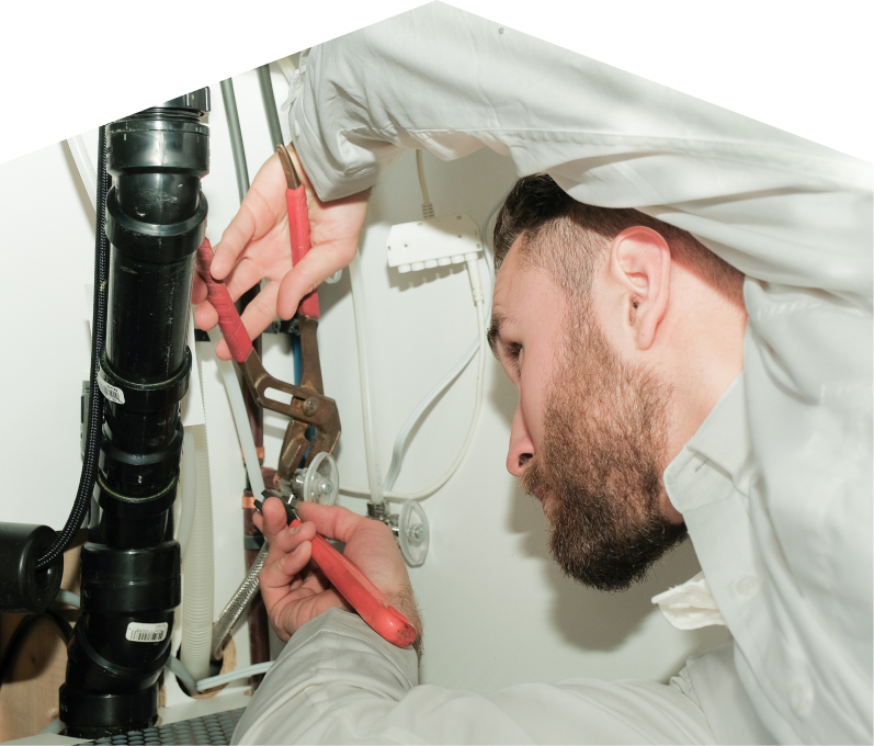 Mersey plumbing specialist uses hand tools to tighten pipe fittings beneath a sink during a repair, with visible wiring and drainage lines in the background. Mersey plumbing specialist uses hand tools to tighten pipe fittings beneath a sink during a repair, with visible wiring and drainage lines in the background.