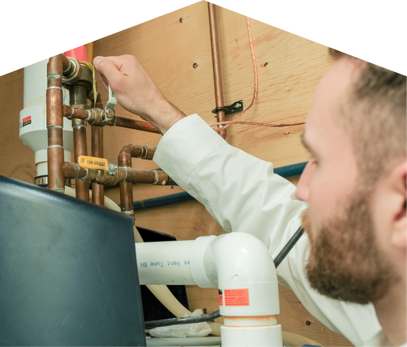 Mersey water heater specialist tightens a valve on copper piping during a water heater installation, with exposed wood paneling and wiring in the background. Mersey water heater specialist tightens a valve on copper piping during a water heater installation, with exposed wood paneling and wiring in the background.